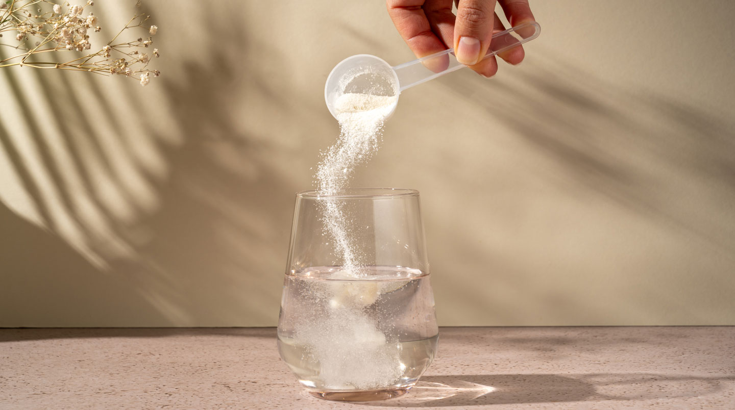 Glass of water with white collagen powder dissolving, measuring spoon on white countertop in natural morning light